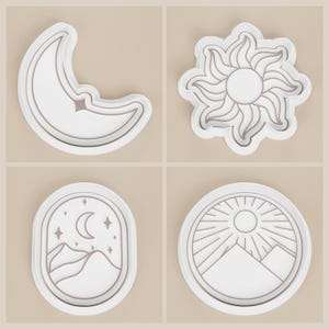 May include: Four white celestial-themed cookie cutters. Designs include a crescent moon with a star, a sunburst, a mountain scene with a moon and stars, and a mountain scene with a sun. The cutters have a detailed outline.