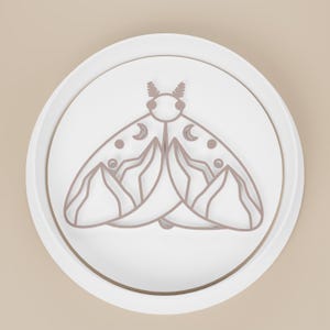May include: A white, circular object with a raised edge, featuring a detailed outline of a moth. The moth design is in a muted brown color and includes crescent moon and dot details on its wings. The object is set against a light beige background.