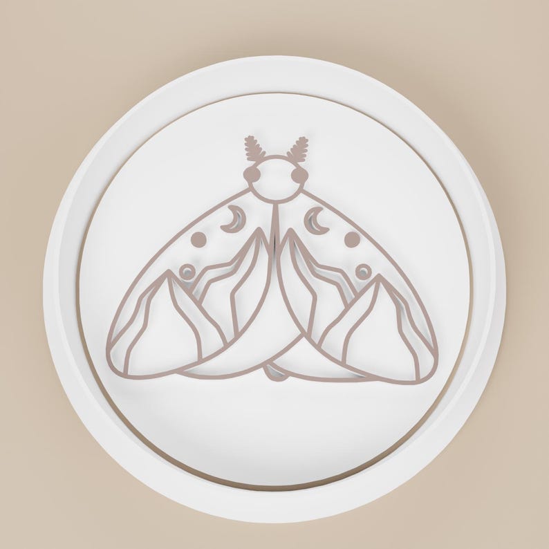 May include: A white, circular object with a raised edge, featuring a detailed outline of a moth. The moth design is in a muted brown color and includes crescent moon and dot details on its wings. The object is set against a light beige background.