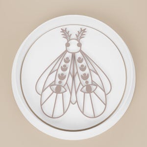 May include: A white, round decorative plate with a raised edge. The plate features a stylized moth design in a muted brown color. The moth has detailed wings, antennae, and eye-like shapes, creating a minimalist, artistic aesthetic.