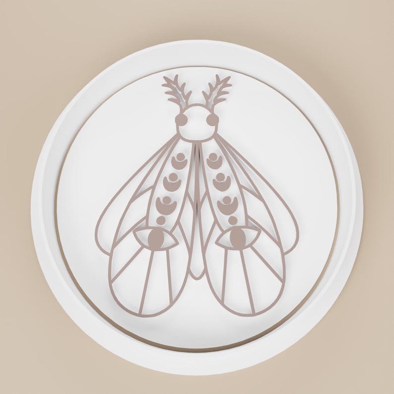 May include: A white, round decorative plate with a raised edge. The plate features a stylized moth design in a muted brown color. The moth has detailed wings, antennae, and eye-like shapes, creating a minimalist, artistic aesthetic.