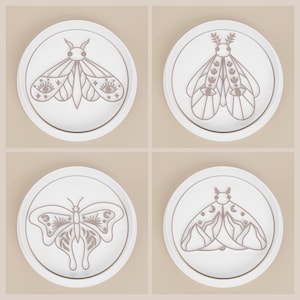 May include: Four white ceramic coasters with a gold rim, each featuring a different moth or butterfly design in shades of brown. The insect illustrations have intricate details, including eyes, moons, and suns.