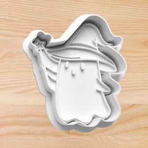 May include: A white plastic cookie cutter in the shape of a cartoon ghost wearing a witch hat and holding a star-tipped wand. The cutter has a layered design and is set against a light wood grain background.