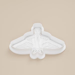 May include: A white plastic cookie cutter in the shape of a moth. The cutter has a detailed wing design and a rounded body. The wings are spread wide, and the overall shape is symmetrical.