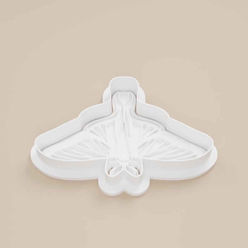 May include: A white plastic cookie cutter in the shape of a moth. The cutter has a detailed wing design and a rounded body. The wings are spread wide, and the overall shape is symmetrical.