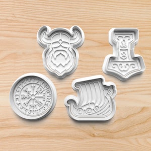 May include: Four white plastic cookie cutters in Viking themes. The shapes include a Viking helmet, Thor's hammer, a Viking ship, and a compass with runes. The cutters are on a light brown wooden surface.