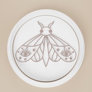May include: A white, circular decorative object featuring a detailed, light brown moth design. The moth has intricate wing patterns with eye and star details. The design is set within a raised, circular border.