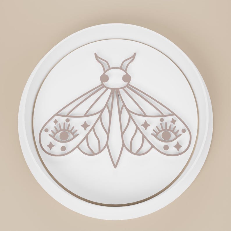 May include: A white, circular decorative object featuring a detailed, light brown moth design. The moth has intricate wing patterns with eye and star details. The design is set within a raised, circular border.