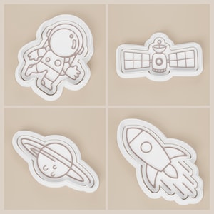May include: Four white cookie cutters with brown outlines, each depicting a space-themed design. The designs include an astronaut, a satellite, a planet with rings, and a rocket ship with flames. The cutters are set against a neutral background.