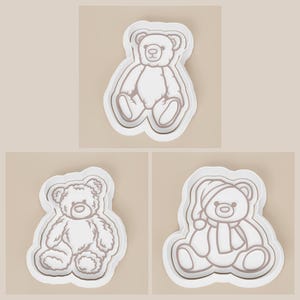 May include: Three white teddy bear-shaped cookie cutters. One bear is sitting upright, another has a textured fur design, and the third is wearing a hat and scarf. The cutters have a white outline and are on a light beige background.