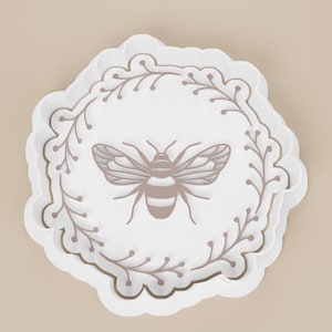 May include: A white, scalloped-edge tray with a bee design. The bee is centered on the tray, surrounded by a brown floral border. The tray is a light, neutral color, suitable for serving or display.