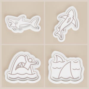 May include: Four white shark-themed cookie cutters with brown outlines. The designs include a shark with open jaws, a shark swimming, a shark attacking a person in the water, and a shark fin in the water.