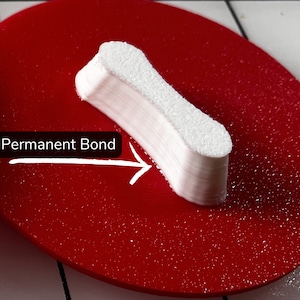 Puede incluir: Un objeto blanco impreso en 3D con una superficie lisa y texturizada. El objeto est&aacute; apoyado sobre una superficie roja con una flecha blanca que apunta hacia &eacute;l y el texto "Permanent Bond".
