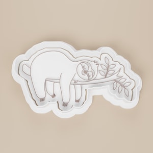 May include: A white plastic cookie cutter in the shape of a sloth resting on a branch with leaves. The sloth is in a relaxed pose, with its eyes closed. The cutter has a detailed outline and is set against a neutral background.