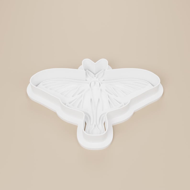May include: A white plastic cookie cutter in the shape of a moth. The cutter has detailed wing and body features. The design is a single piece with a rounded edge and a heart-shaped head.
