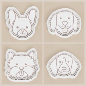 May include: Four white dog-shaped cookie cutters. The cutters feature the faces of different dog breeds, including a Corgi, a Golden Retriever, a West Highland White Terrier, and a Beagle. Each cutter has a detailed outline.