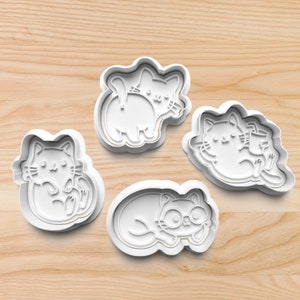 May include: Four white plastic cat-shaped cookie cutters in various poses. One cat is curled up, another is lying down with glasses, and two others are in playful positions. The cutters are on a light wood surface.
