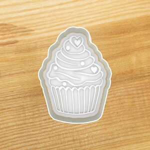 May include: White plastic cookie cutter shaped like a cupcake with frosting and hearts.