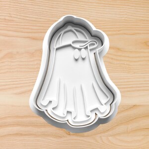 May include: White plastic cookie cutter shaped like a ghost wearing a baseball cap.