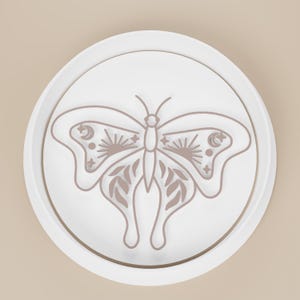 May include: A white, circular decorative object featuring a detailed butterfly design. The butterfly has intricate wing patterns with sun, moon, and star motifs. The design is in a muted brown color, set against a white background.