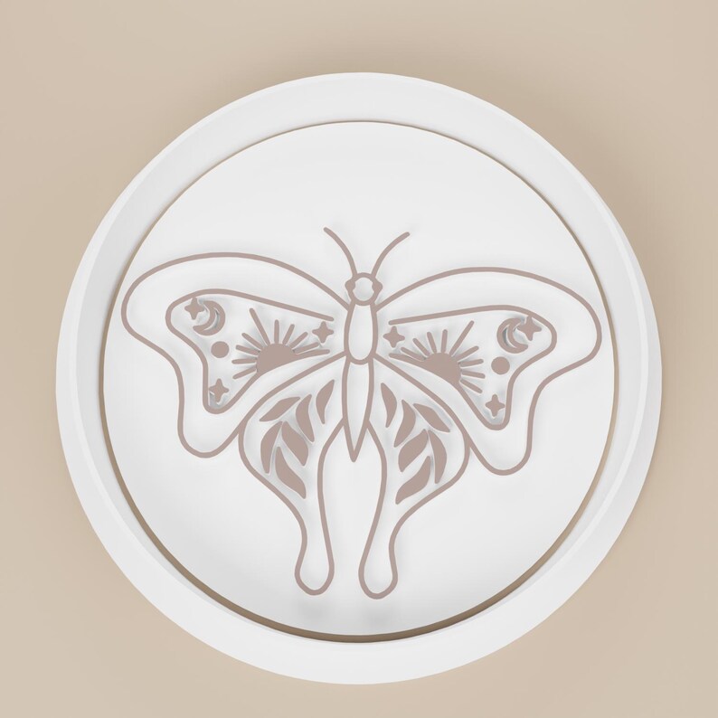 May include: A white, circular decorative object featuring a detailed butterfly design. The butterfly has intricate wing patterns with sun, moon, and star motifs. The design is in a muted brown color, set against a white background.
