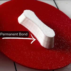 May include: A white, rectangular object with a textured surface, labeled "Permanent Bond" with an arrow, is placed on a red, circular surface. The object appears to be made of stacked layers, suggesting a bonding or adhesive product.