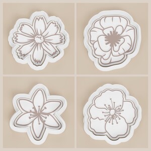 May include: Four white flower-shaped cookie cutters with brown outlines. Each cutter has a different floral design, including a daisy, poppy, and other stylized blossoms. The cutters are set against a neutral background.