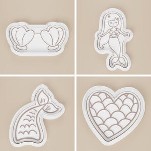 May include: Four white plastic cookie cutters in mermaid theme. The shapes include a pair of seashells, a mermaid, a mermaid tail, and a heart with a scale pattern. The cutters have a raised outline.