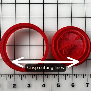 May include: Two red plastic cookie cutters. One is a circle, and the other is a round shape with a clock design. The image shows the phrase "Crisp cutting lines". The cookie cutters are on a grid background.