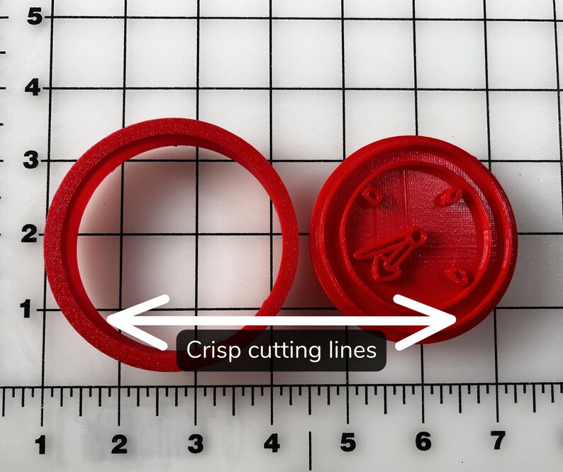 May include: Two red plastic cookie cutters. One is a circle, and the other is a round shape with a clock design. The image shows the phrase "Crisp cutting lines". The cookie cutters are on a grid background.
