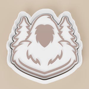 May include: A white and taupe cookie cutter in the shape of a yeti with two pine trees on either side. The yeti has a heart-shaped face and a furry body. The cookie cutter has a layered design.