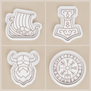 May include: Four white and tan cookie cutters with Viking-themed designs. The designs include a longboat, Thor's hammer, a Viking helmet, and a compass rose. The cutters are made of plastic and are suitable for baking.