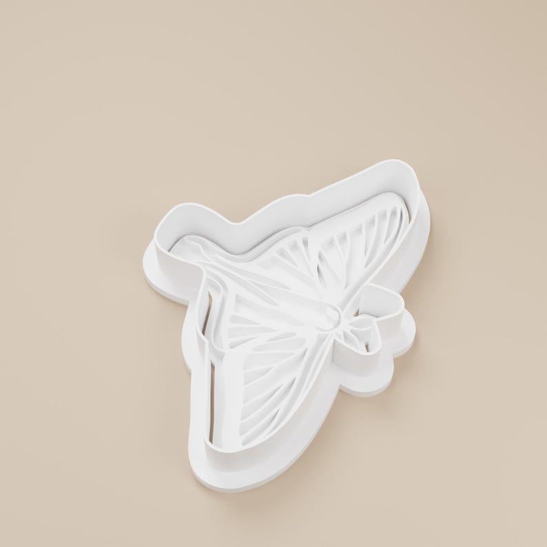 May include: A white plastic butterfly-shaped cookie cutter. The cutter has detailed wing patterns and a rounded edge. The design is suitable for baking and crafting projects. The cutter is on a light beige background.