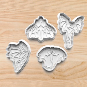 May include: Four white plastic cookie cutters in various dragon designs. The cutters include a dragon with outstretched wings, a dragon with a long tail, and two other dragon-themed shapes. The cutters are on a light wood surface.