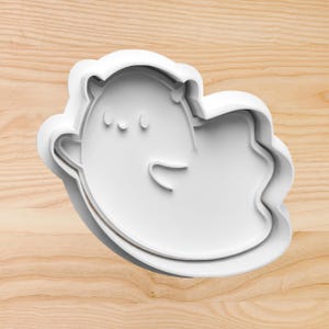 May include: White plastic cookie cutter shaped like a cartoon ghost with a cute expression. The ghost has two eyes and a small mouth.