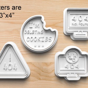 May include: Four white plastic cookie cutters shaped like a computer screen, a triangle with an exclamation point, a cookie with a bite taken out of it, and a computer screen with a face. The computer screens have the text "404 Not Found" and "404" on them. The triangle has the text "404" on it. The cookie has the text "I'M DELETING COOKIES" on it. The cutters are approximately 3 inches by 4 inches.
