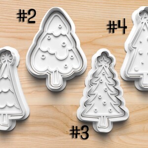 May include: Four white plastic Christmas tree-shaped cookie cutters. Each cutter has a different design, including a star on top. The numbers #1, #2, #3, and #4 are printed above or next to each cutter.