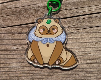 May include: A clear acrylic keychain featuring a cartoon raccoon wearing a blue and white scarf with a green leaf on its head. The keychain has a green metal clasp.