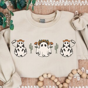 May include: A beige sweatshirt with a graphic print of three ghosts wearing cowboy hats and holding lassos. The ghosts are surrounded by cacti and sunflowers.