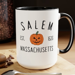 May include: White ceramic mug with a black rim and handle. The mug features a black and orange illustration of a jack-o'-lantern with the text "SALEM EST. 1626 MASSACHUSETTS".