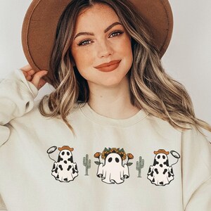 May include: A cream-colored sweatshirt with a graphic of three ghosts wearing cowboy hats and holding lassoes. The ghosts are in front of cacti and one ghost has a flower crown.