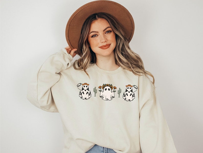 May include: A cream-colored sweatshirt with a graphic of three ghosts wearing cowboy hats and holding lassoes. The ghosts are in front of cacti and one ghost has a flower crown.