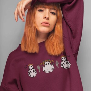 May include: A maroon sweatshirt with a graphic of three ghosts wearing cowboy hats and floral crowns. Two of the ghosts are wearing cow print and one is a traditional white ghost. The ghosts are surrounded by cacti.