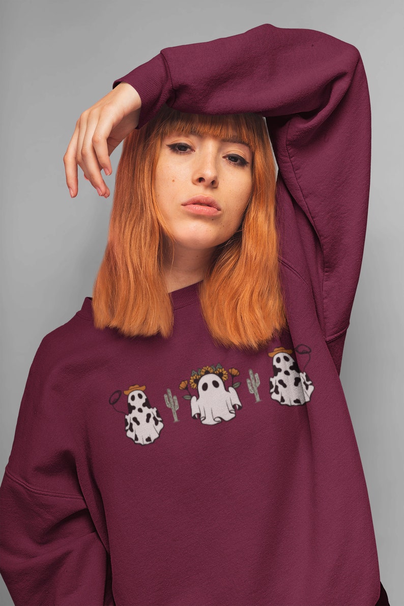 May include: A maroon sweatshirt with a graphic of three ghosts wearing cowboy hats and floral crowns. Two of the ghosts are wearing cow print and one is a traditional white ghost. The ghosts are surrounded by cacti.