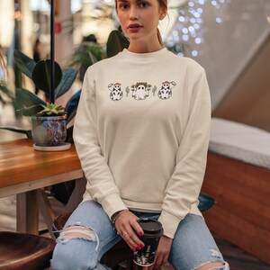 May include: A woman wearing a cream colored sweatshirt with a graphic of three ghost cows wearing cowboy hats and holding a cup of coffee. The sweatshirt has long sleeves and a crew neck.