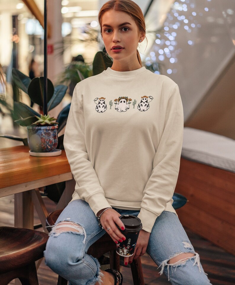 May include: A woman wearing a cream colored sweatshirt with a graphic of three ghost cows wearing cowboy hats and holding a cup of coffee. The sweatshirt has long sleeves and a crew neck.