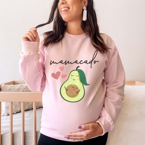 May include: Pink sweatshirt with the text "mamacado" and a cartoon avocado with a smaller avocado inside. The avocado has a smiling face and closed eyes. There are three pink hearts above the avocado.