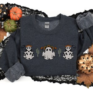 May include: A dark gray sweatshirt with a graphic of three ghosts wearing cowboy hats and holding lassoes. The ghosts are surrounded by cacti and sunflowers.