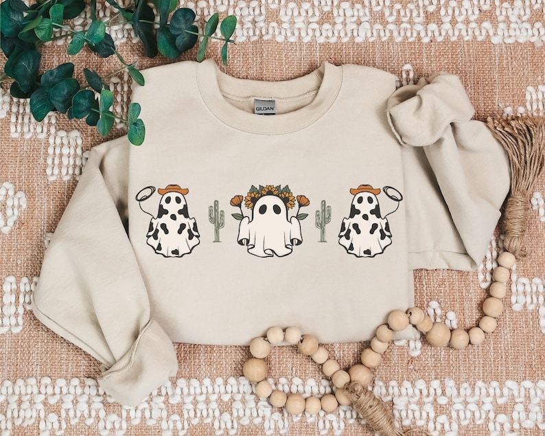 May include: A beige sweatshirt with a graphic print of three ghosts wearing cowboy hats and holding lassos. The ghosts are surrounded by cacti and sunflowers.