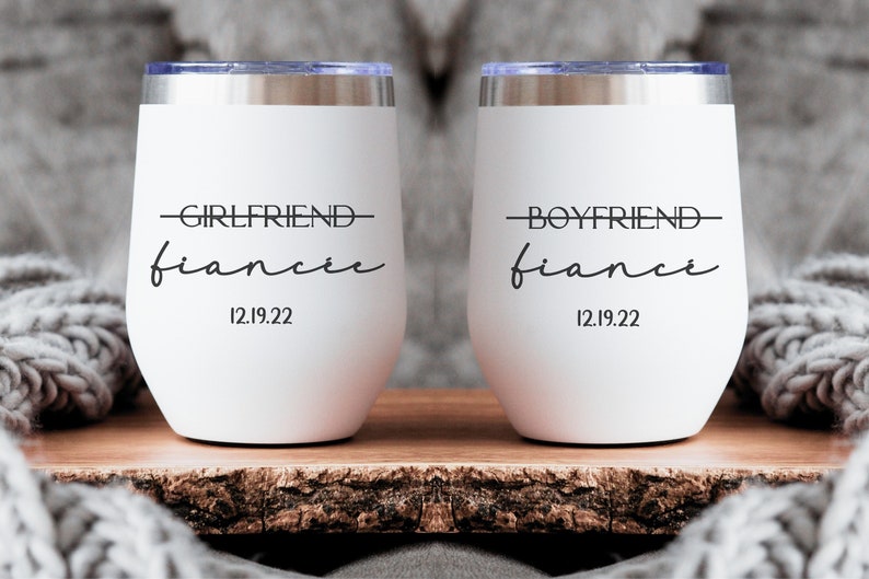May include: Two white stainless steel wine tumblers with black text. The left tumbler says "Girlfriend fiance 12.19.22" and the right tumbler says "Boyfriend fiance 12.19.22".